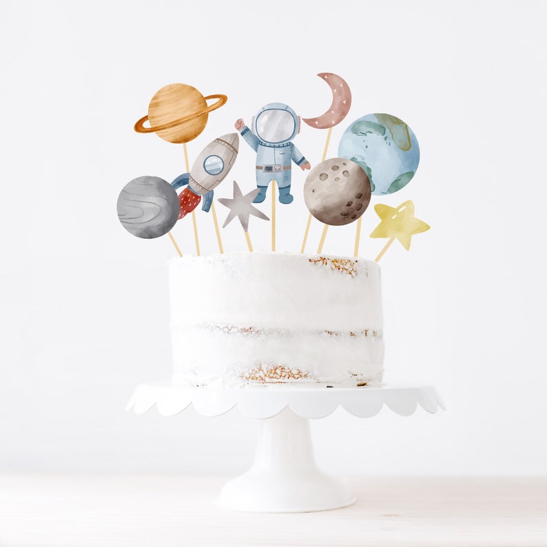 Pode incluir: Um bolo branco com uma variedade de toppers de bolo com tema espacial, incluindo um astronauta, um foguete, planetas, uma lua e uma estrela. O bolo est&aacute; em um suporte de bolo branco.