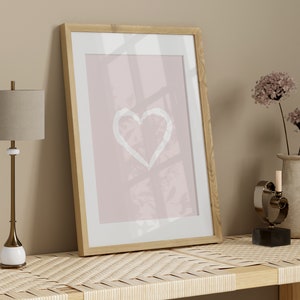 May include: A framed print with a white heart shape on a pink background, a lamp with a beige lampshade, a vase with dried flowers, and a metal heart sculpture on a woven shelf.