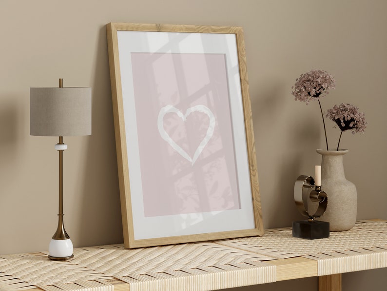May include: A framed print with a white heart shape on a pink background, a lamp with a beige lampshade, a vase with dried flowers, and a metal heart sculpture on a woven shelf.