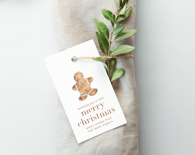 May include: A white gift tag with a watercolor gingerbread man illustration and the text "wishing you a very merry christmas". The tag is tied with twine to a linen napkin and decorated with green leaves.