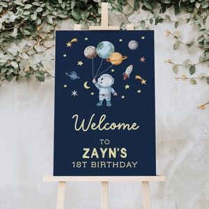 Può includere: Un cartello di benvenuto blu con un astronauta dei cartoni animati che tiene palloncini a forma di pianeti. Il cartello dice "Benvenuto al 1° compleanno di Zayn".