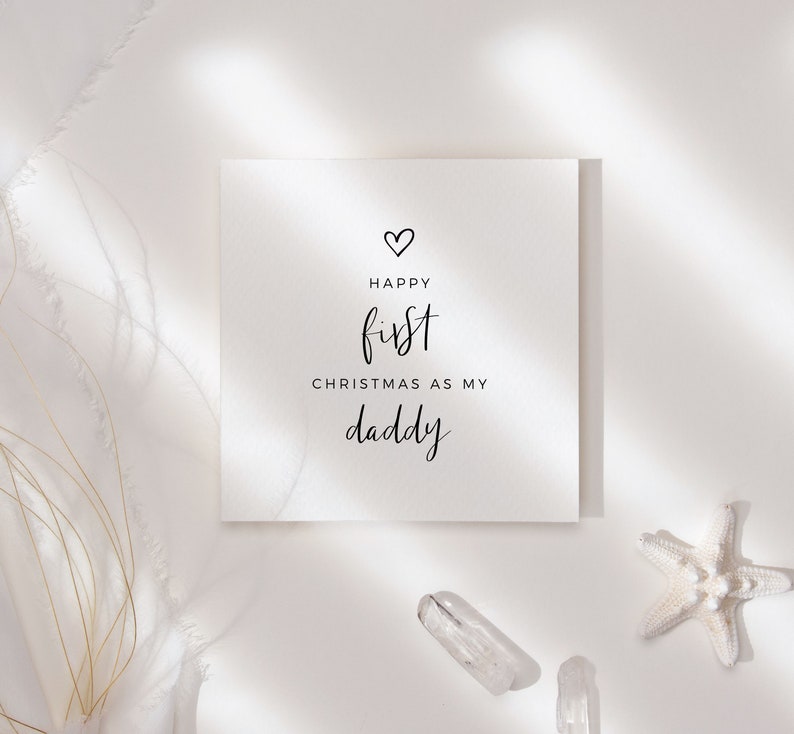 May include: A white card with black text that reads "Happy first Christmas as my daddy" with a small heart above the word "Happy".