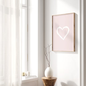 May include: A pink framed print with a white heart drawn on it. The print is hanging on a white wall next to a window with white sheer curtains. A white vase with a brown branch is on a wooden stool.