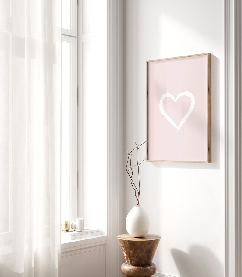 May include: A pink framed print with a white heart drawn on it. The print is hanging on a white wall next to a window with white sheer curtains. A white vase with a brown branch is on a wooden stool.
