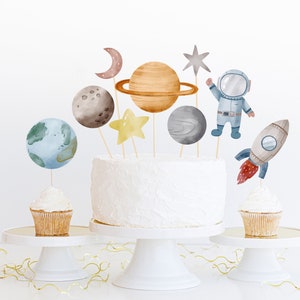Pode incluir: Um bolo branco decorado com toppers de astronauta, foguete, estrela e planeta. O bolo est&aacute; em um suporte de bolo branco. H&aacute; dois cupcakes com toppers combinando em suportes de bolo brancos.