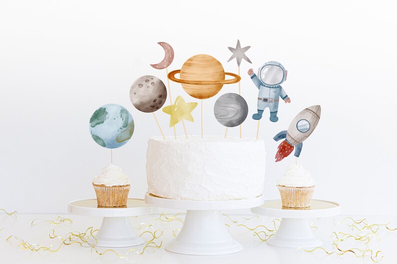 Pode incluir: Um bolo branco decorado com toppers de astronauta, foguete, estrela e planeta. O bolo est&aacute; em um suporte de bolo branco. H&aacute; dois cupcakes com toppers combinando em suportes de bolo brancos.