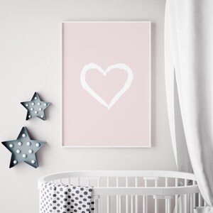 May include: A white framed poster with a pink background and a white hand-drawn heart in the center. The poster is hanging on a white wall above a white crib with a black and white polka dot sheet and blanket.