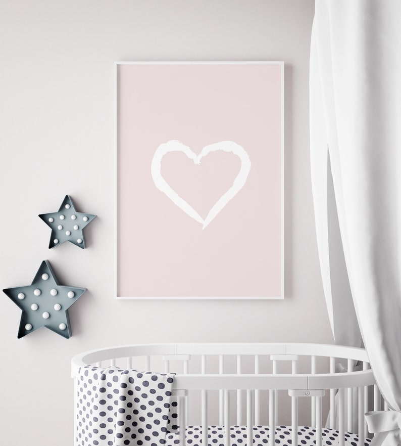 May include: A white framed poster with a pink background and a white hand-drawn heart in the center. The poster is hanging on a white wall above a white crib with a black and white polka dot sheet and blanket.