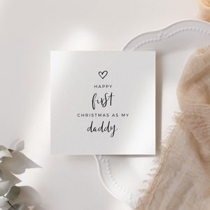 May include: A white card with black text that reads "Happy first Christmas as my daddy" with a small heart above the word "Happy".
