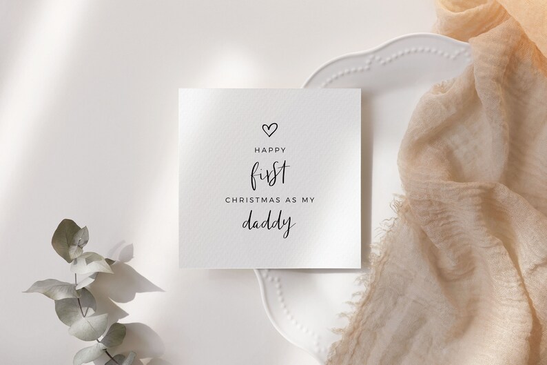 May include: A white card with black text that reads "Happy first Christmas as my daddy" with a small heart above the word "Happy".