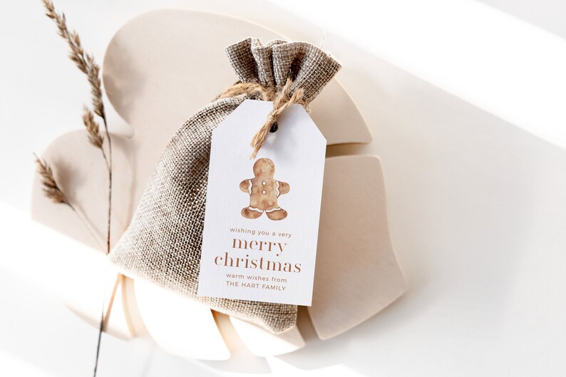 May include: A small burlap gift bag with a tag featuring a watercolor gingerbread man. The tag reads "wishing you a very merry christmas" and "warm wishes from The Hart Family". The bag is tied with twine.