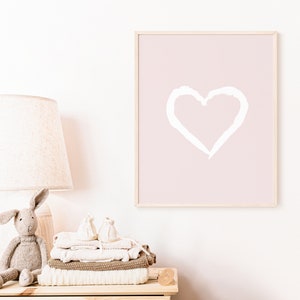 May include: A framed print with a white heart shape on a light pink background. The print is hanging on a white wall above a wooden dresser with a lamp and a stack of folded blankets.