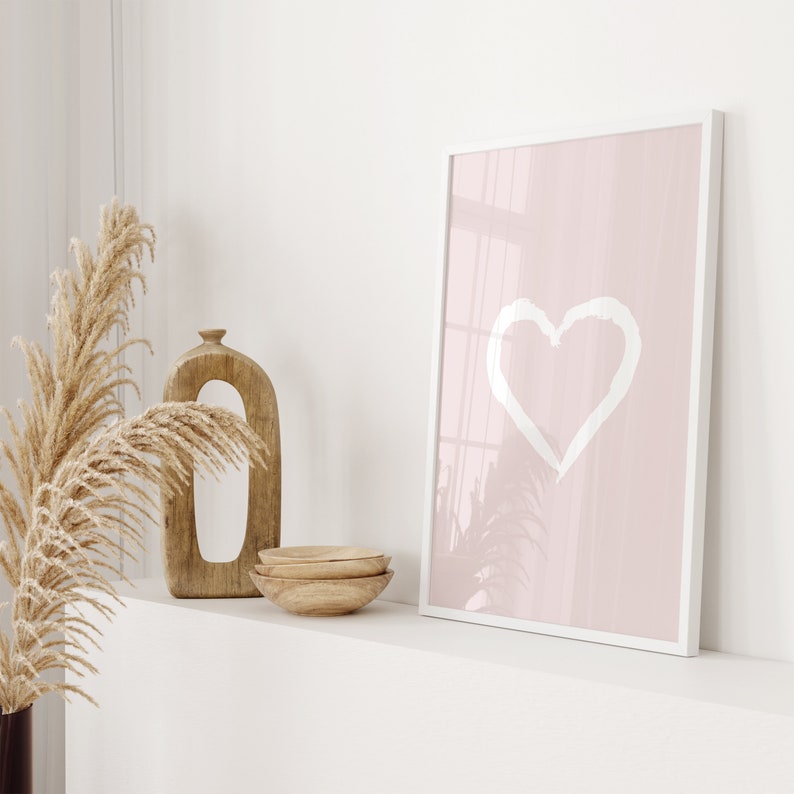 May include: A white framed print with a pink background and a white heart shape in the center. The print is sitting on a white shelf with a wooden vase and bowls.