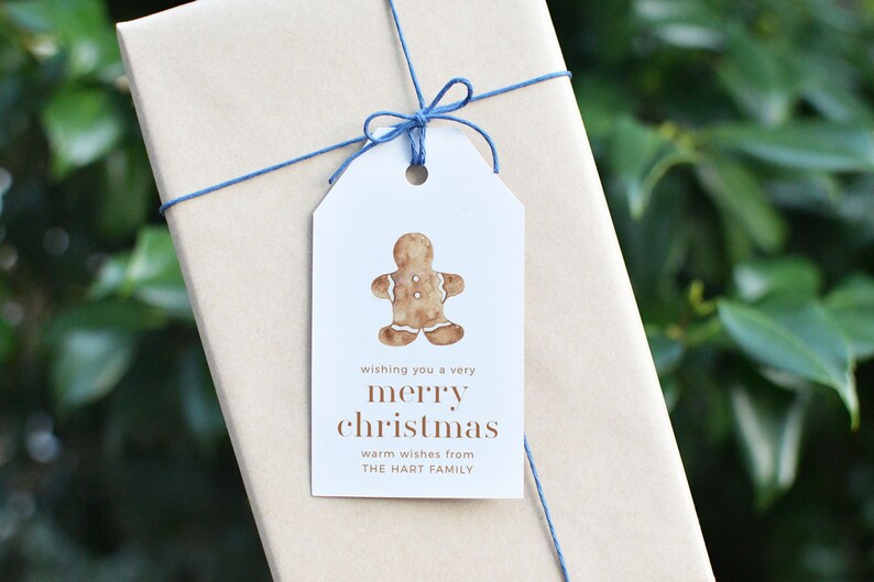 May include: A wrapped gift with a white tag featuring a watercolor gingerbread man. The tag reads "wishing you a very merry christmas" and "warm wishes from THE HART FAMILY". The gift is tied with blue string.