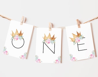 May include: A banner with the word "ONE" in black letters, each letter on a separate card. Each card has a gold crown with pink and blue flowers on top.