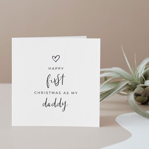 May include: A white card with black text that reads "Happy first Christmas as my daddy" with a small heart symbol above the word "Happy".