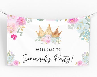 May include: A white banner with a gold crown and floral accents in pink, blue, and green. The text "WELCOME TO Savannah's Party!" is written in black script. The banner is suspended by two wires.