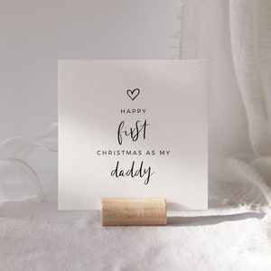 May include: White card with black text that reads "Happy first Christmas as my daddy" with a small heart above the word "Happy". The card is standing on a wooden stand.