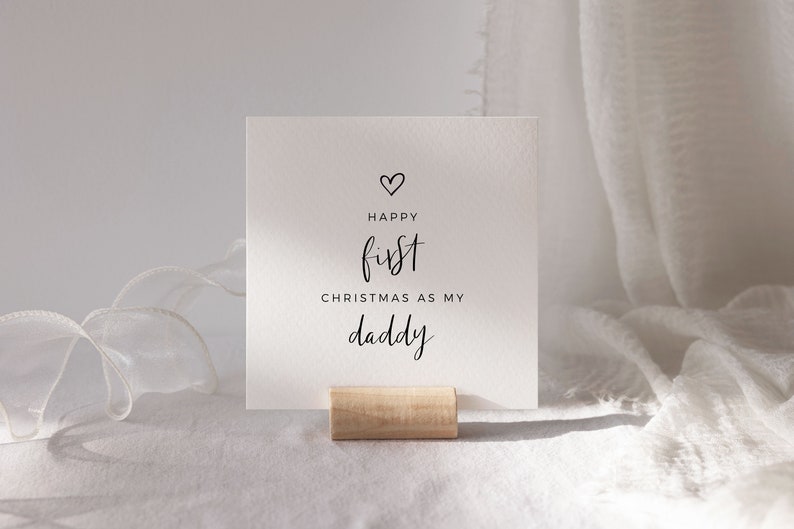 May include: White card with black text that reads "Happy first Christmas as my daddy" with a small heart above the word "Happy". The card is standing on a wooden stand.