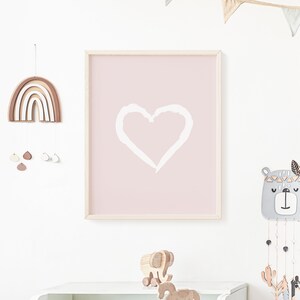 May include: A pink framed print with a white heart shape on a light pink background. The print is hanging on a white wall with a wooden rainbow mobile and a wooden bear mobile hanging above a white dresser. The dresser has a wooden rocking horse and a wooden elephant toy on top.