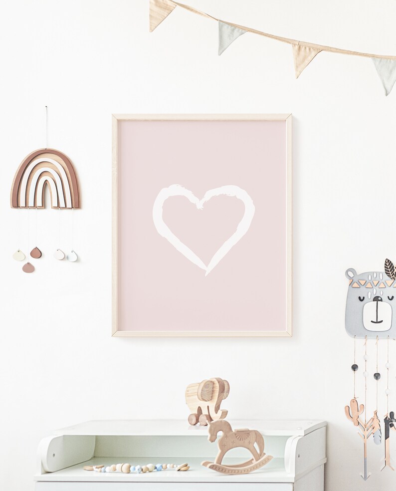 May include: A pink framed print with a white heart shape on a light pink background. The print is hanging on a white wall with a wooden rainbow mobile and a wooden bear mobile hanging above a white dresser. The dresser has a wooden rocking horse and a wooden elephant toy on top.