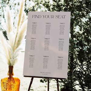 May include: A white sign with the text "Find Your Seat" and a seating chart for a wedding reception. The sign is on an easel in a grassy outdoor setting.