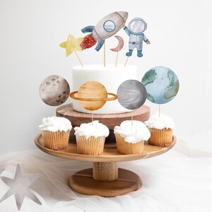 Pode incluir: Um bolo branco decorado com toppers de astronauta, foguete e planetas. O bolo &eacute; cercado por cupcakes com toppers combinando.