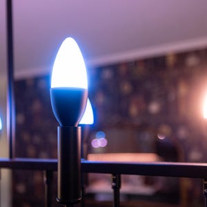 May include: A modern chandelier featuring several lit, white, teardrop-shaped LED bulbs. The bulbs are mounted on black metal rods, creating a sleek design. The background is blurred, with hints of a patterned wall.