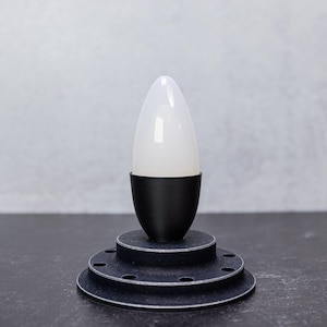May include: A modern black and white lamp with a conical white bulb. The lamp sits on a tiered, circular base with a matte finish. The lamp is on a dark surface against a neutral background.