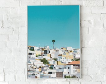 Puede incluir: Una impresión enmarcada que presenta un pueblo costero bajo un cielo azul brillante. Edificios blancos con detalles amarillos y naranjas se agrupan, con una sola palmera en el centro. La impresión se muestra contra una pared de ladrillos blancos.