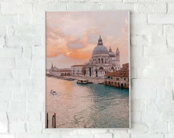 May include: A photograph of a large, white church with a dome, located on a canal in Venice, Italy. The sky is a soft pink and orange, suggesting a sunset. A small boat is visible in the water.