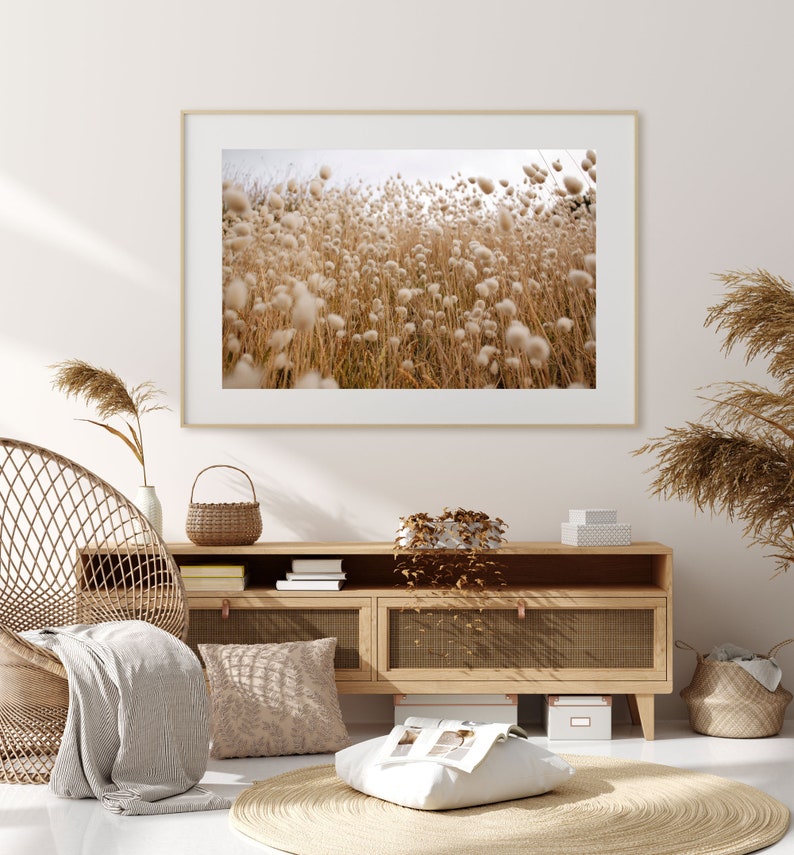 May include: A framed photograph of a field of tall, fluffy, white grasses blowing in the wind. The photo is hanging on a white wall above a wooden cabinet with a woven basket and books on top.