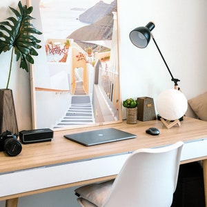 May include: A white desk with a wooden top and a white drawer. A laptop computer, a camera, a speaker, a lamp, a potted plant, and a moon lamp are on the desk. A white chair with wooden legs is in front of the desk. A framed print of a white building with a staircase leading up to a doorway is on the wall behind the desk.