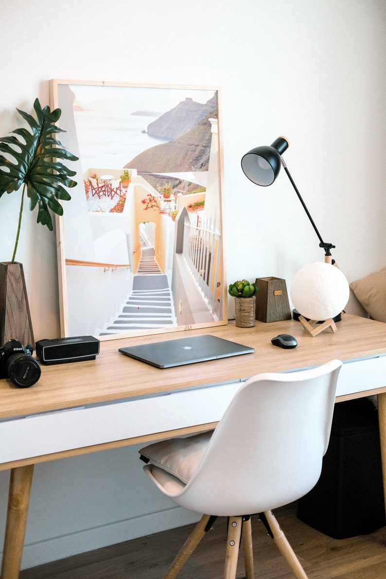 May include: A white desk with a wooden top and a white drawer. A laptop computer, a camera, a speaker, a lamp, a potted plant, and a moon lamp are on the desk. A white chair with wooden legs is in front of the desk. A framed print of a white building with a staircase leading up to a doorway is on the wall behind the desk.