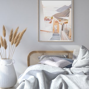 May include: A white bed with a wooden headboard and a white blanket. The bed is in a room with a white wall and a framed photograph of a white building with a staircase leading down to the ocean.