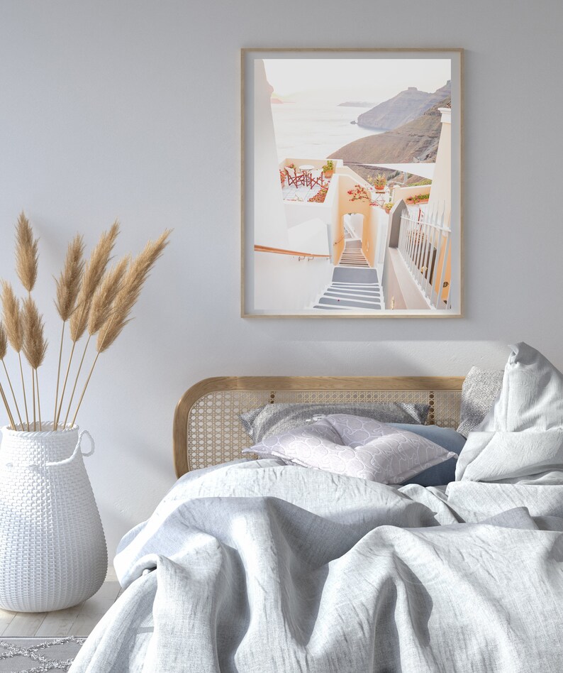 May include: A white bed with a wooden headboard and a white blanket. The bed is in a room with a white wall and a framed photograph of a white building with a staircase leading down to the ocean.