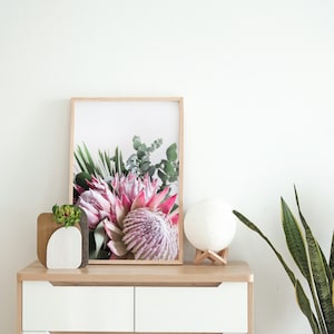 以下が含まれることがあります： 白い背景にピンクと緑のプロテアの花が描かれた額装されたプリント。プリントは、引き出しが2つ付いた白いドレッサーの上に置かれています。ドレッサーの上には、白いプランターに入った小さな多肉植物があります。白いムーンランプがドレッサーの上に置かれています。画像の右側には、白い鉢に入った緑のヘビの植物があります。