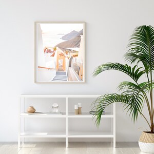 May include: A framed photograph of a white building with a staircase leading down to a blue ocean. The building is surrounded by white walls and a green hillside. The photo is hanging on a white wall above a white shelf with decorative items.