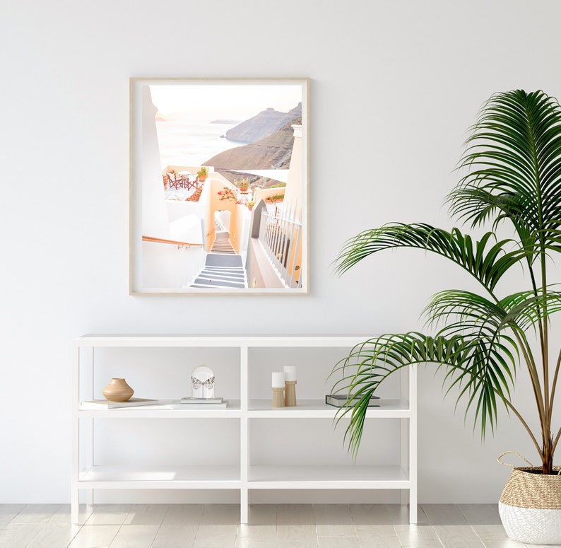 May include: A framed photograph of a white building with a staircase leading down to a blue ocean. The building is surrounded by white walls and a green hillside. The photo is hanging on a white wall above a white shelf with decorative items.