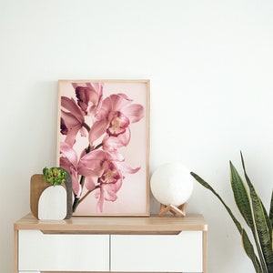 May include: A framed print of a pink orchid flower against a light pink background. The print is on a wooden dresser with a white moon lamp and a potted snake plant.