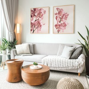 May include: A living room with a light grey sofa, two framed prints of pink orchids, a wooden coffee table, a wooden side table, and a knitted pouf. The room has a light grey rug and a large window with a sheer curtain.