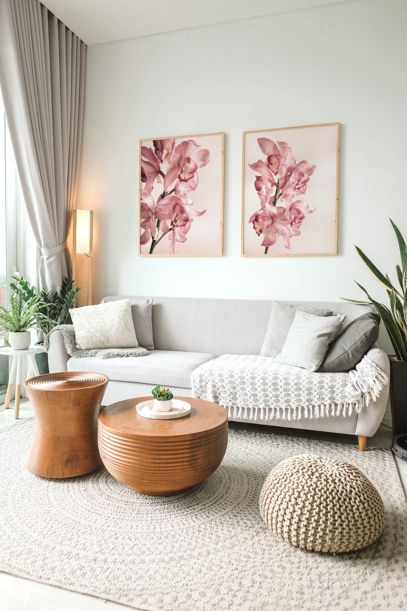 May include: A living room with a light grey sofa, two framed prints of pink orchids, a wooden coffee table, a wooden side table, and a knitted pouf. The room has a light grey rug and a large window with a sheer curtain.