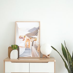 May include: A white dresser with two drawers and wooden handles. A framed photograph of a white building with stairs leading down to a blue ocean is on top of the dresser. A white moon lamp and a potted plant are also on the dresser.