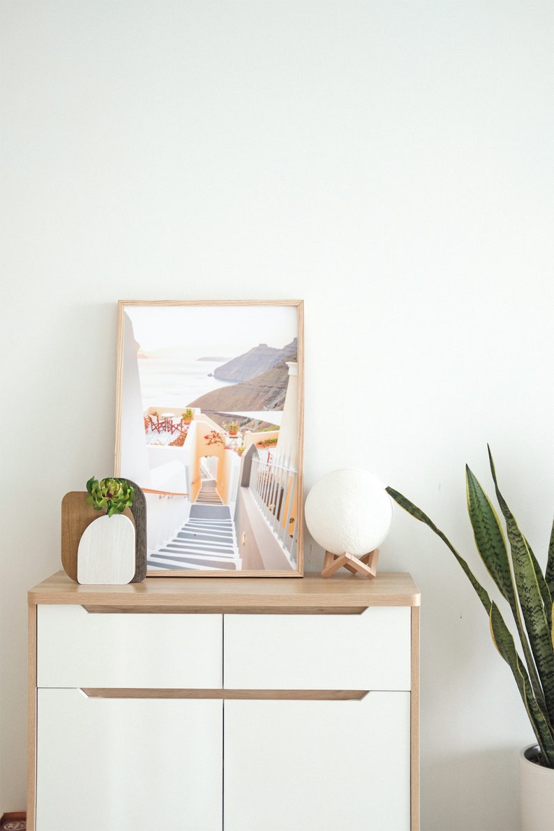May include: A white dresser with two drawers and wooden handles. A framed photograph of a white building with stairs leading down to a blue ocean is on top of the dresser. A white moon lamp and a potted plant are also on the dresser.