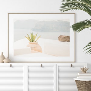 May include: A framed photograph of a white building with a potted plant and a rock in the foreground. The building is set against a backdrop of a hazy, blue ocean and mountains.