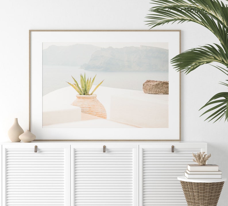May include: A framed photograph of a white building with a potted plant and a rock in the foreground. The building is set against a backdrop of a hazy, blue ocean and mountains.