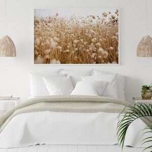 May include: A white bed with a beige throw blanket and white pillows in a bedroom with a framed photograph of a field of tall grass with white fluffy seed heads. There are two hanging light fixtures with woven shades and a potted plant on a side table.