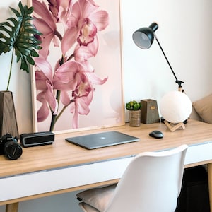 May include: A light wood desk with a white surface and a white chair. A laptop computer, a speaker, a camera, and a lamp are on the desk. A framed print of pink orchids is on the wall behind the desk.