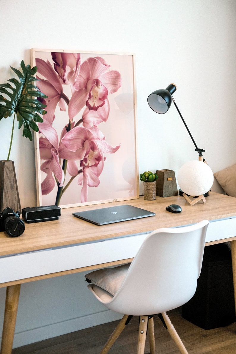 May include: A light wood desk with a white surface and a white chair. A laptop computer, a speaker, a camera, and a lamp are on the desk. A framed print of pink orchids is on the wall behind the desk.