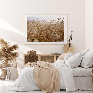 May include: A white bed with a tan blanket and white pillows in a bedroom with a framed photograph of a field of dried flowers and a wooden cabinet with a wicker basket.